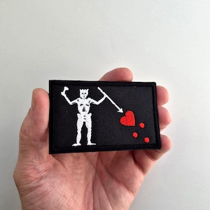 May include: Black rectangular patch featuring a white skeleton figure holding a spear, with a red heart and three red dots. The patch is held in a hand against a white background. The design is embroidered.
