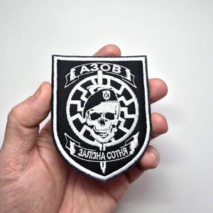 May include: Black and white embroidered patch featuring a skull wearing a beret, surrounded by a circular design. The patch has the word "AZOV" at the top and "ZALIZNA SOTNYA" at the bottom. The patch is held in a hand.