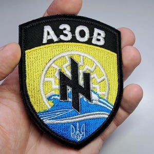 Puede incluir: Parche bordado en negro, amarillo y azul con la palabra "АЗОВ" en blanco. El parche presenta un emblema estilizado con un diseño similar al sol y un símbolo de tridente.