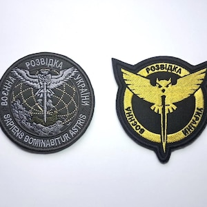 Op de afbeelding: Twee geborduurde patches. Eén is rond met een uil en zwaard ontwerp, omringd door tekst. De andere is schildvormig met een gouden uil en zwaard ontwerp op een zwarte achtergrond, met tekst. Militaire insignes.