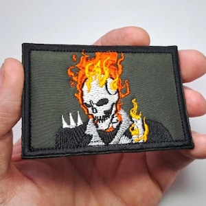 May include: Embroidered patch featuring a flaming skull design. The skull is white with orange and yellow flames. The patch has a black border and is set against a dark green background. The patch is rectangular.
