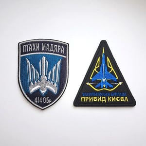 Op de afbeelding: Twee geborduurde patches. Eén is schildvormig met een blauw en wit ontwerp en Oekraïense tekst. De andere is een zwarte driehoek met een blauwe jet en gele accenten, ook met Oekraïense tekst.
