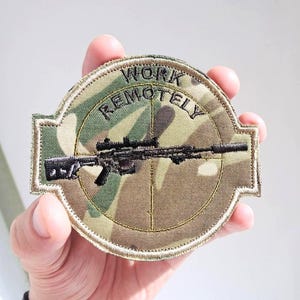 Pode incluir: Um patch de camuflagem com as palavras "WORK REMOTELY" arqueadas sobre um rifle preto com mira telescópica. O patch, segurado em uma mão, tem um design circular com um esquema de cores marrom e verde.