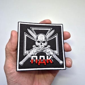 May include: Square black and white patch featuring a skull, crossed swords, and a white cross design. The patch has the red letters "ПДК" and a white border. The patch is held in a hand.