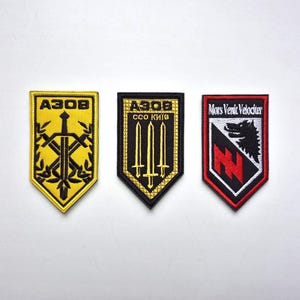 Puede incluir: Tres parches bordados con forma de escudo. El primero es amarillo con una espada y el texto "A3OB". El segundo es negro con tres espadas y el texto "A3OB CCO Kиiв". El tercero es rojo, negro y blanco con un lobo y el texto "Mors Venit Velociter".