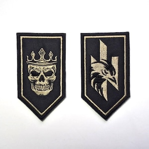Puede incluir: Dos parches negros bordados. Uno presenta una calavera con corona, el otro una cabeza de águila. Ambos están delineados con hilo dorado y tienen la parte inferior puntiaguda. Los parches están sobre un fondo blanco.