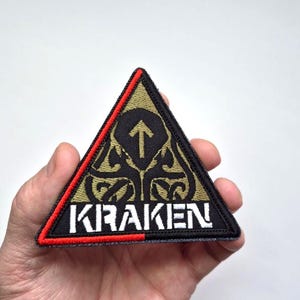Può includere: Una toppa triangolare con la scritta "KRAKEN" in lettere bianche. La toppa ha uno sfondo nero con un disegno oro e nero e un bordo rosso. Una freccia punta verso l'alto all'interno del disegno.