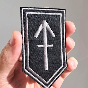 May include: Black and white embroidered patch with a shield shape. The patch features a stylized arrow symbol in the center, with a white border. The patch is held in a hand.
