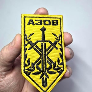 Könnte beinhalten: Gelb-schwarzer gestickter Aufnäher mit dem Wort "A3OB" oben. Der Aufnäher zeigt ein schwarzes Schwert und andere Symbole. Der Aufnäher wird in einer Hand gehalten.