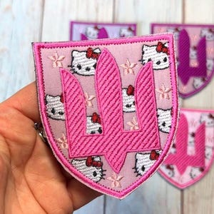 May include: A pink embroidered patch in a shield shape. The patch features a pink trident design and Hello Kitty faces with red bows. The background is light pink with small flower designs. Other patches are visible in the background.