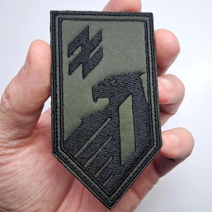 May include: Olive green embroidered patch with a black eagle and a stylized symbol. The patch is rectangular with a pointed base and a black border. The design is detailed and appears to be machine-embroidered.