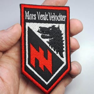 Może przedstawiać: Haftowana naszywka z czarnym tłem, czerwoną obwódką i białymi akcentami. Naszywka zawiera napis "Mors Venit Velociter", głowę wilka i czerwony zygzak. Naszywka jest trzymana w dłoni.