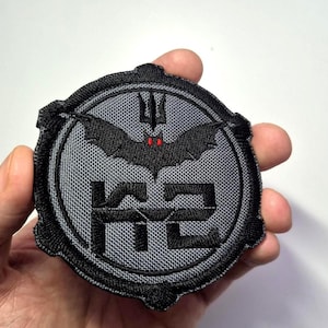 May include: Round, gray and black embroidered patch featuring a bat with red eyes above the letters "K2". The patch has a textured background and a black border. The bat has a trident above it.