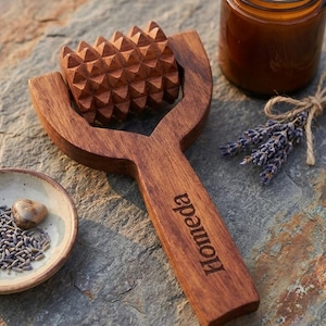 Könnte beinhalten: Eine Holz-Massagerolle mit strukturierter Oberfläche, neben einer brennenden Kerze in einem Glas, einer kleinen Schale mit Lavendel und einem Bündel getrocknetem Lavendel. Auf der Rolle steht das Wort "Homeda".