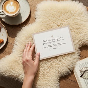 Puede incluir: Una escena acogedora con un capuchino con arte latte, un cruasán y una tarjeta que dice "gracias por tu compra". La tarjeta está sobre una alfombra de piel de oveja color crema.
