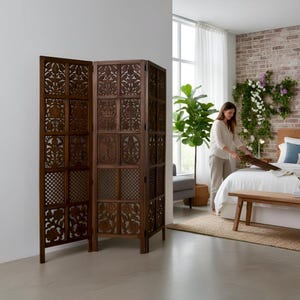 May include: A dark brown, three-panel room divider with intricate floral and geometric cut-out designs. The wooden screen stands in a well-lit room, adding a decorative element to the space. The screen is a functional and stylish home decor item.