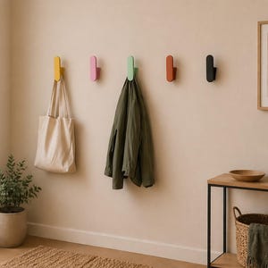 Puede incluir: Un perchero de pared con cinco ganchos de colores: amarillo, rosa, verde, naranja y negro. Una bolsa de lona cuelga del gancho amarillo y una chaqueta verde cuelga del gancho verde. También se ven una impresión enmarcada y una mesa pequeña.