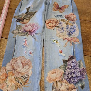May include: Light blue denim jeans featuring distressed areas and colorful printed graphics of birds, butterflies, ants, insects, and various floral arrangements including peonies and lilacs. A pink ruler is visible on the left side of the jeans.