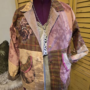 May include: A patchwork jacket featuring a mix of olive green plaid, mauve bleached fabric, and floral rose prints. The jacket has a collared neckline, long sleeves with cuffs, two rounded front pockets with pink trim, and three brown buttons.