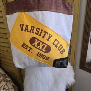 May include: White canvas tote bag with brown trim and handles. A large yellow patch with "VARSITY CLUB XXL EST. 1969" in brown lettering is sewn on the front. The bag has a rectangular shape and is designed for carrying items.