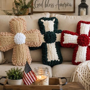 May include: Three cross-shaped pillows made of chunky, textured yarn. The left pillow is tan with a white center, the middle pillow is dark green with a white center, and the right pillow is red with a white center, all featuring a plush, looped finish.