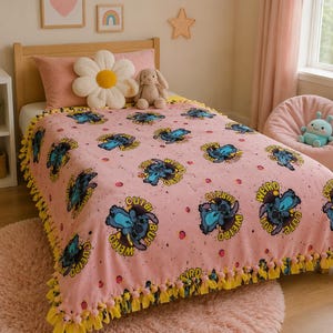 May include: Pink throw blanket featuring a repeating pattern of a blue alien character with the words "Cute But Weird". The blanket has a yellow fringe trim and is on a bed with a pink pillow and a stuffed animal.