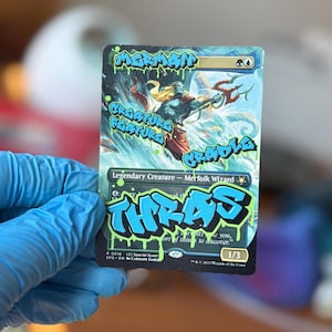 May include: A collectible card featuring a Merfolk Wizard illustration, overlaid with graffiti-style text in blue and green. The card includes the text "MERMAN", "CREATURE FEATURE", "CRADLE", and "THRAS". The card is held by a person wearing a blue glove.