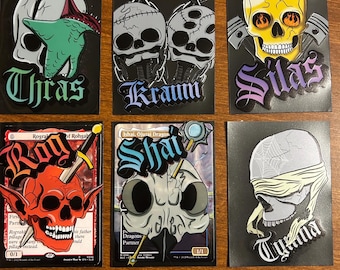 May include: Six black cards with skull illustrations. The cards feature various designs, including a skull with a green hand, two chained skulls, a yellow skull with pistons, a red skull with horns, a skull with a magnifying glass, and a skull with a blindfold. Each card has a name.
