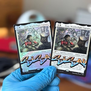 May include: Two "Sisay, Weatherlight Captain" trading cards are held in a gloved hand. The cards feature a colorful illustration of a woman and other figures. The text on the cards includes "Legendary Creature - Human Soldier" and other game-related text.