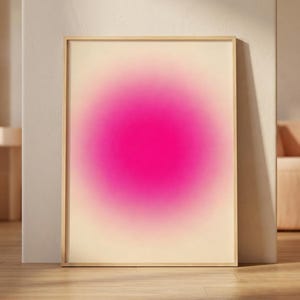 May include: A framed art print featuring a vibrant pink gradient circle against a soft, off-white background. The artwork is encased in a light-colored wooden frame, creating a modern aesthetic. The piece is displayed indoors, leaning against a wall.