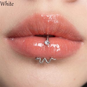 Pode incluir: Um piercing labial de tom prateado com um pendente em forma de cobra na parte inferior e uma pedra redonda transparente e facetada na parte superior. A joia está posicionada verticalmente através do lábio inferior.