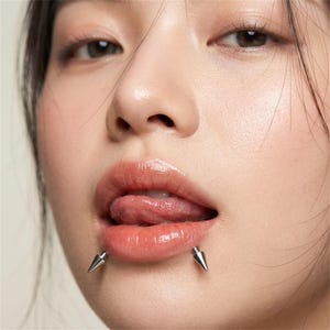 May include: Close-up of a person's face with lip piercings. Two silver, cone-shaped piercings are visible on the lower lip. The person's tongue is extended, and the lips are glossy and a natural pink color.