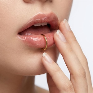 May include: A smooth, polished gold-tone circular hoop ring positioned on the lower lip. The metal ring features a simple, continuous band design with a reflective surface, creating a clean and understated appearance against the skin.