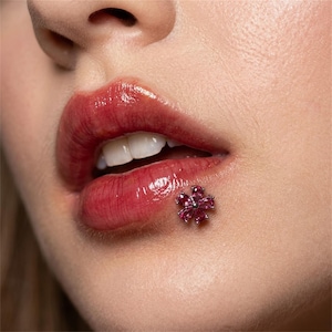 Può includere: Primo piano di labbra con rossetto rosso lucido e un piccolo piercing a forma di fiore. Il piercing presenta più pietre preziose rosa incastonate in un metallo argentato. Le labbra sono leggermente socchiuse.
