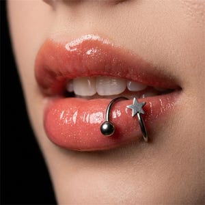 Puede incluir: Un aro labial en espiral de tono plateado con una pequeña bola en un extremo y una estrella de cinco puntas en el otro. La joya está colocada en el labio inferior, cubierto con un producto labial brillante de tono coral.