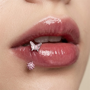 Op de afbeelding: Een zilveren lip piercing met een vlinderbedel met lichtroze emaille vleugels op de bovenlip, gecombineerd met een aparte onderlip piercing bestaande uit een kleine, ronde, met roze kristallen ingelegde knop in zilverkleurig metaal.
