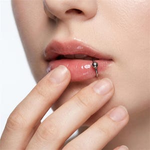 Puede incluir: Un aro labial circular de tono plateado que presenta un cierre de bola lisa individual. La joya está colocada en el labio inferior, creando una estética metálica y limpia. El diseño de aro circular es sencillo, pulido y de aspecto brillante.