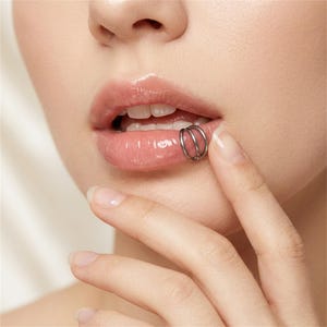 Puede incluir: Un aro de labio de triple argolla en tono plateado que presenta tres bandas circulares finas e interconectadas. La joya está colocada en el labio inferior, mostrando un acabado metálico y un diseño continuo en capas que rodea el borde del labio.
