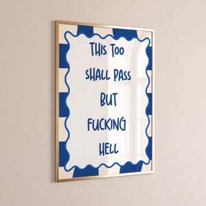 以下が含まれることがあります： 青い文字で「THIS TOO SHALL PASS BUT FUCKING HELL」と書かれた額入りのアートプリント。アートワークは、青い波状のボーダーと金色のフレームが付いた白い背景です。背景には青と白のチェック柄があります。