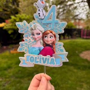 Może przedstawiać: Ozdoba na tort z Elsą i Anną z Krainy Lodu, z cyfrą 4 i imieniem "OLIVIA". Ozdoba zawiera błyszczący wz&oacute;r płatka śniegu i małą figurkę Olafa. Postacie są w swoich charakterystycznych strojach.
