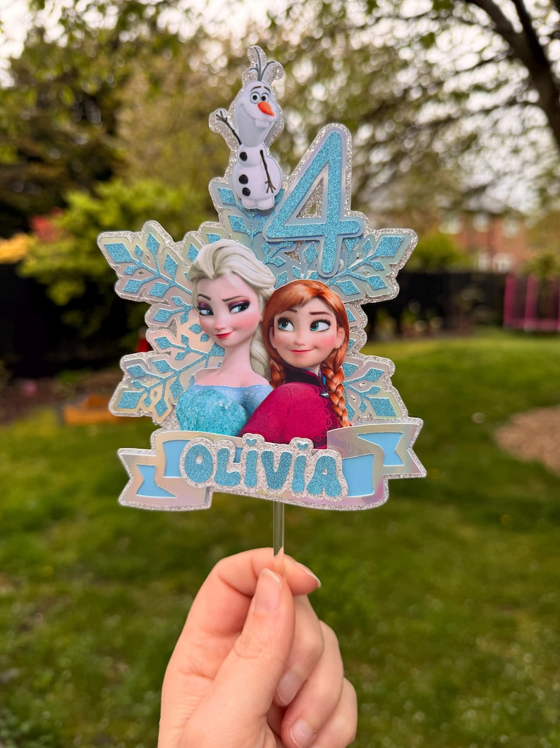 Może przedstawiać: Ozdoba na tort z Elsą i Anną z Krainy Lodu, z cyfrą 4 i imieniem "OLIVIA". Ozdoba zawiera błyszczący wz&oacute;r płatka śniegu i małą figurkę Olafa. Postacie są w swoich charakterystycznych strojach.