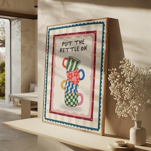May include: A framed rectangular wall art print featuring a cream and beige checkered background with a scalloped blue border. The centre displays the text "PUT THE KETTLE ON" above a vertical stack of four colourful mugs with striped, floral, and checkered patterns.