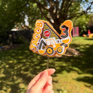 May include: A colorful cake topper with a construction theme. The topper features a yellow and black road sign with the word "STOP", a construction vehicle, the number "3", and the name "JACOB".