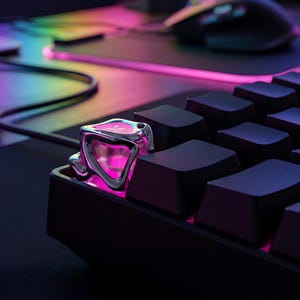 Puede incluir: Primer plano de un teclado negro con una tecla plateada personalizada. La tecla tiene un diseño único con detalles en rosa y plata, iluminada por un brillo rosa. El fondo incluye un ratón y una superficie iluminada colorida.