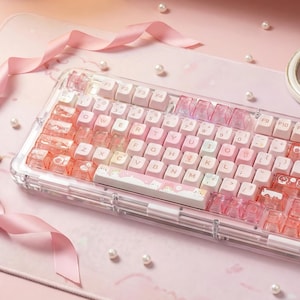 May include: A clear, mechanical keyboard with pink and white keycaps. The keyboard has a clear frame and a pink base. The keycaps are a mix of white, pink, and clear, with some featuring decorative designs. Pink ribbons and pearls surround the keyboard.
