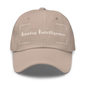May include: A beige baseball cap with the text "Analog Intelligence" in white. The text is enclosed in a white rectangular frame. The cap has a curved brim and a button on top.
