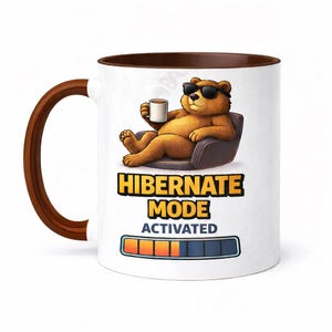 Puede incluir: Taza de cerámica blanca con interior y asa marrón. La taza presenta un oso de dibujos animados con gafas de sol y sosteniendo una taza de café, tumbado en una silla. El texto en la taza dice "HIBERNATE MODE ACTIVATED" con una barra de progreso.