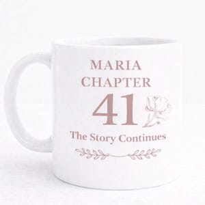 Puede incluir: Taza de cerámica blanca con asa curva. La taza presenta el texto "MARIA CHAPTER 41 The Story Continues" en color rosa suave, junto con una ilustración floral y ramitas decorativas.