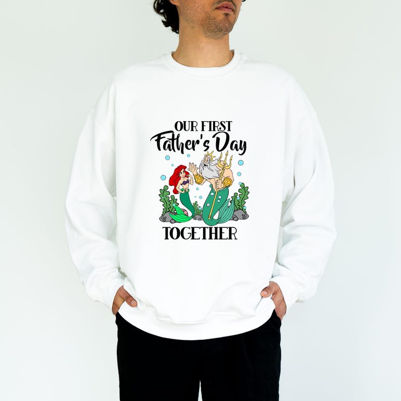 Puede incluir: Sudadera blanca con un gr&aacute;fico que presenta una sirena y un trit&oacute;n, con el texto "OUR FIRST Father's Day TOGETHER". La ilustraci&oacute;n incluye a la sirena con cabello rojo y al trit&oacute;n con un tridente. La sudadera es de estilo casual.
