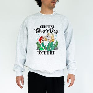 Puede incluir: Una sudadera gris claro con el texto "OUR FIRST Father's Day TOGETHER" en escritura negra. El gr&aacute;fico presenta una sirena y un trit&oacute;n de dibujos animados. La sirena tiene el pelo rojo y una cola verde. El trit&oacute;n tiene barba blanca y cola verde.